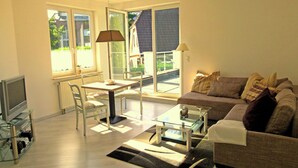 A modern living room with a sofa, a glass coffee table, a dining table with chairs, a TV, and large windows offering a view of the outside. - Vacation apartments on the Lesum - FeWo Lesmona - TOP long-term conditions (Bremen)