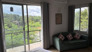 A room with a bed, a green sofa with patterned cushions, and large windows offering a view of greenery and a house.