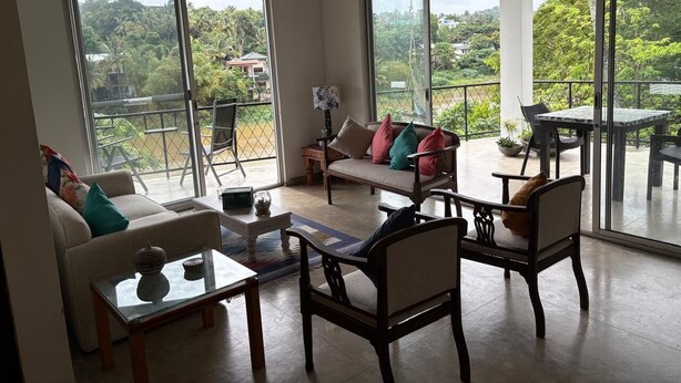 A spacious living room with a sofa, armchairs, and a coffee table. Large sliding glass doors lead to an outdoor patio with a table and chairs.