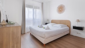A minimalist bedroom with a large bed, bedside tables, a hanging light fixture, and a view through sheer curtains.