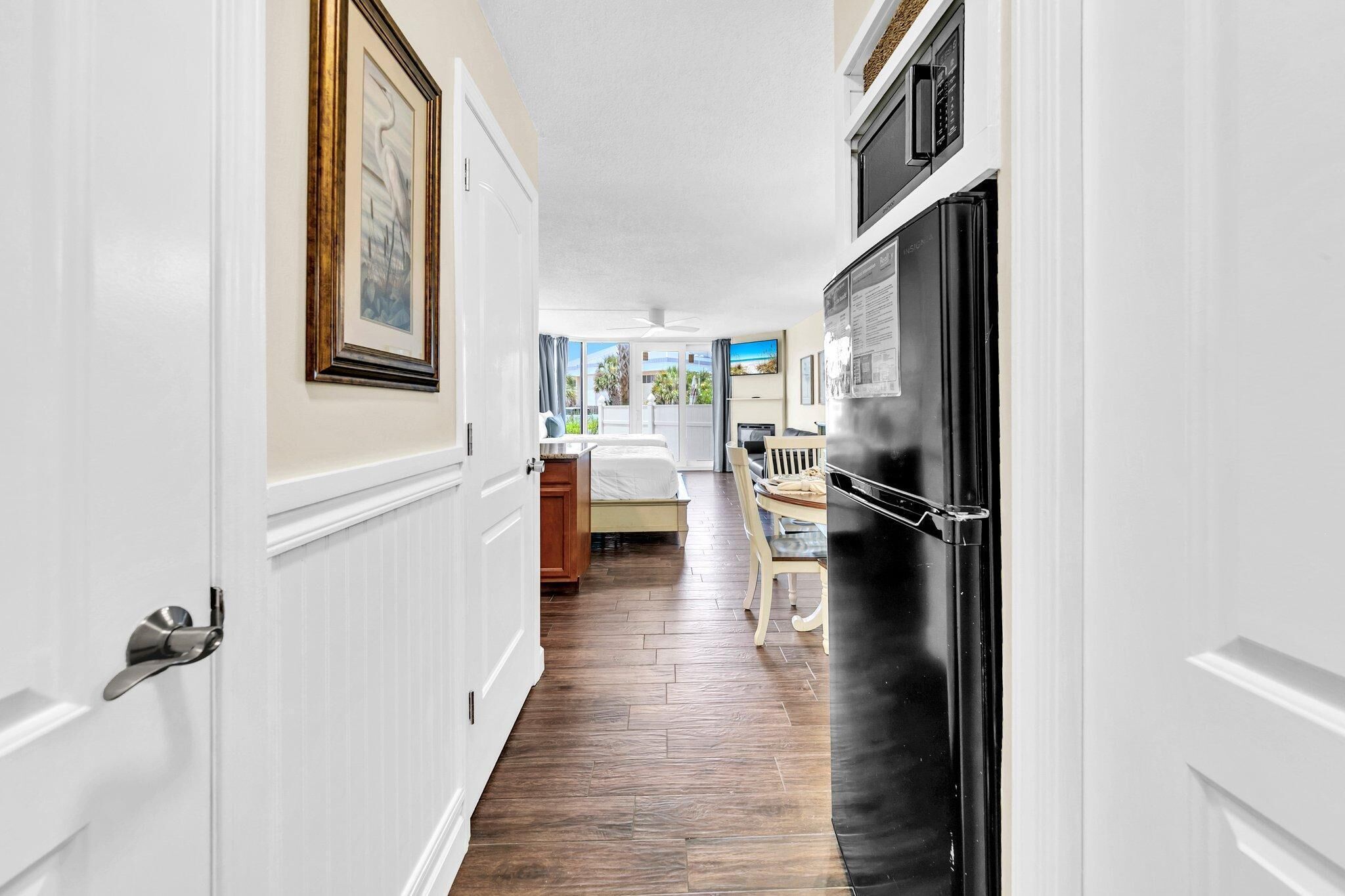 A narrow hallway with a wooden floor, white walls, and a black refrigerator. There is a bed with a white headboard and a window with a view of the outdoors.