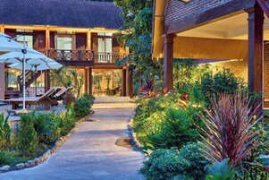 Un chemin bien entretenu traverse un jardin et mène à un bâtiment doté d’un balcon, le tout étant entouré d’une végétation luxuriante et de plantes.
