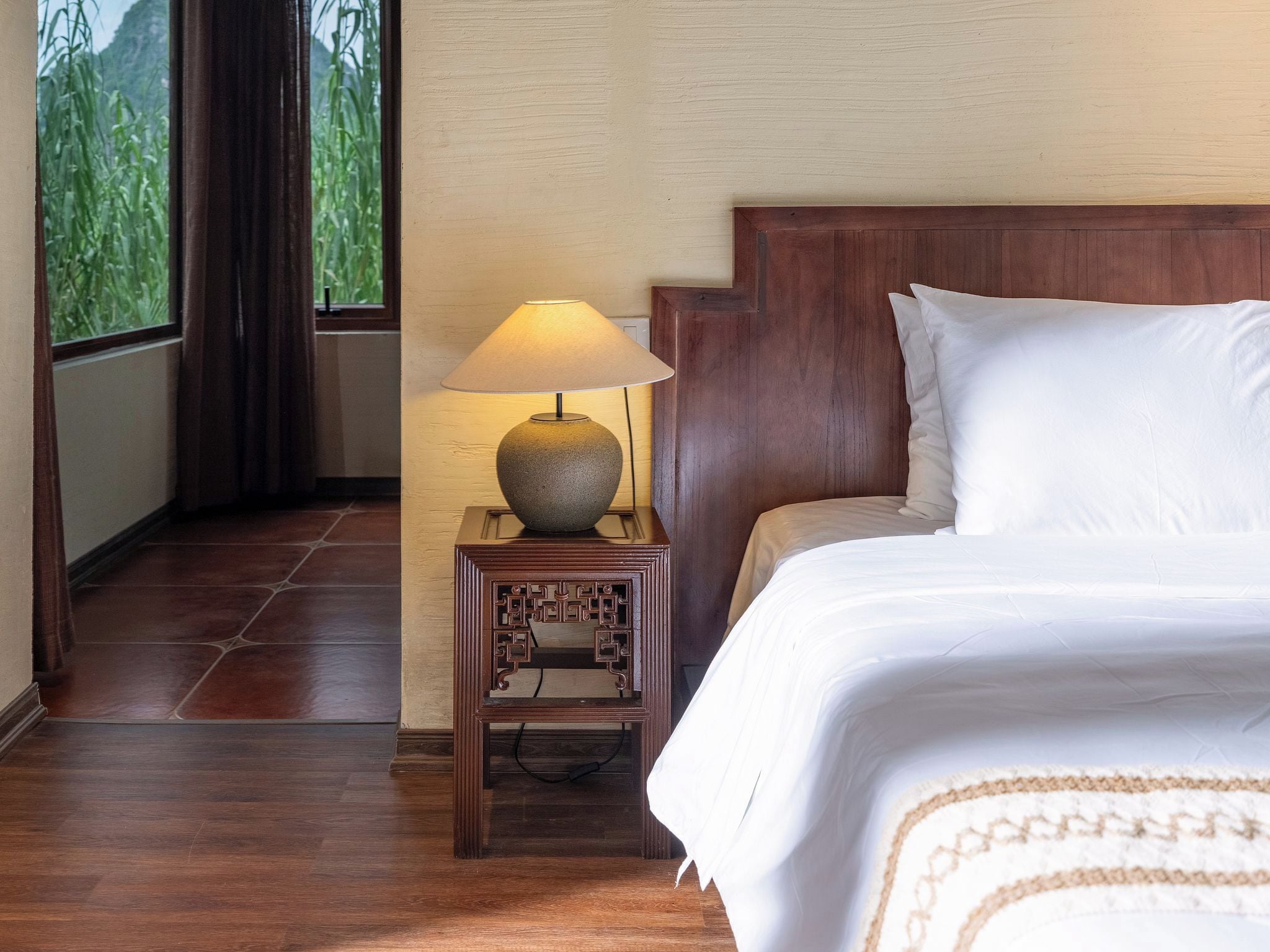 A hotel room with a wooden headboard, a bed with white linens, a small table with a lamp, and a view of greenery through a window.