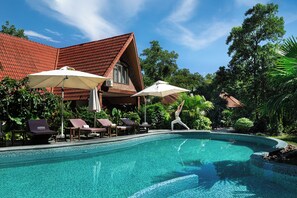 Une piscine entourée de chaises longues et de parasols, elle-même bordée d’une végétation luxuriante et d’un bâtiment doté d’un toit en tuiles.