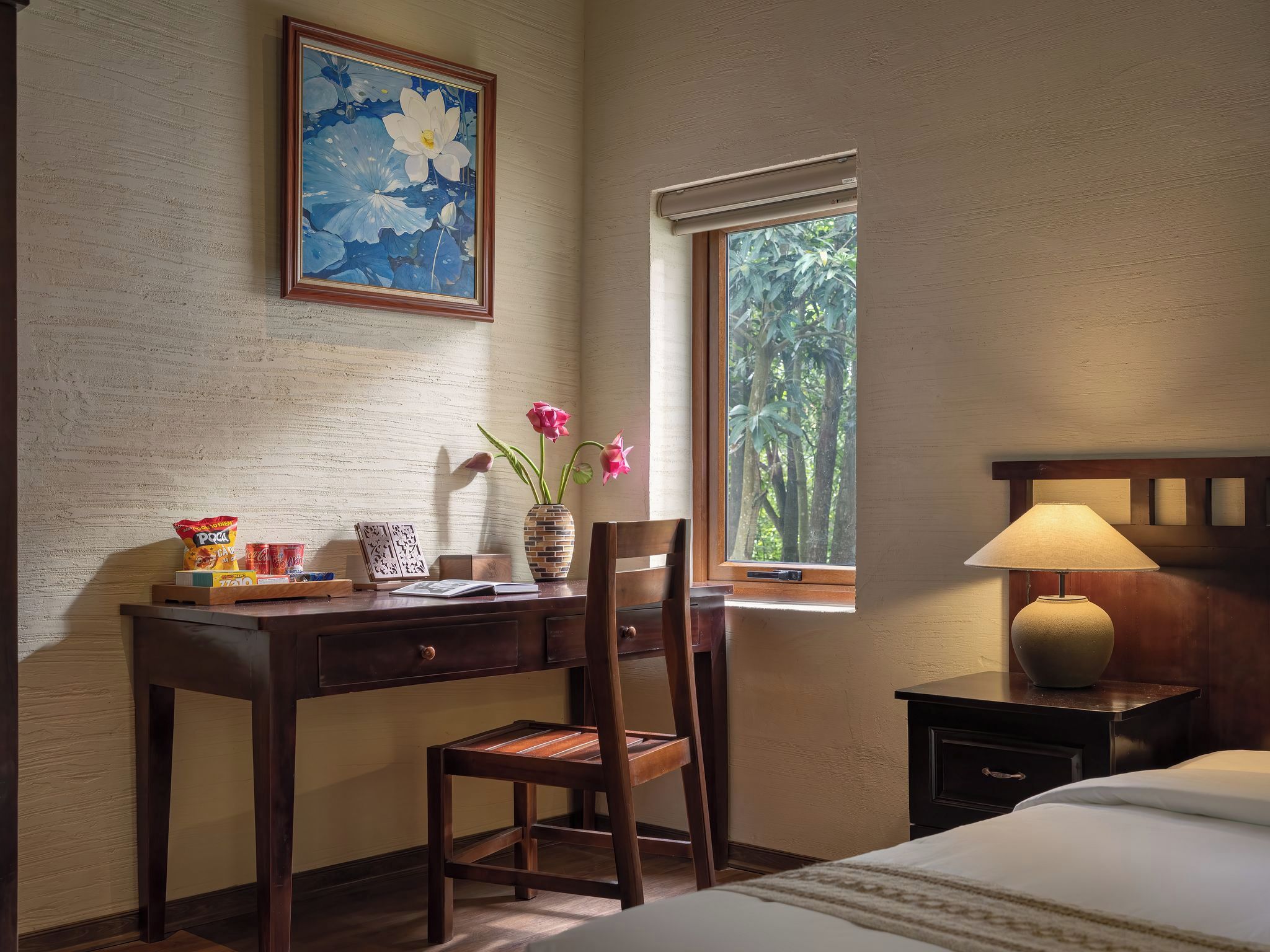 Una habitación con un escritorio de madera, una silla y una mesita de noche con lámpara. Se ve una ventana con vistas a árboles y un cuadro en la pared.