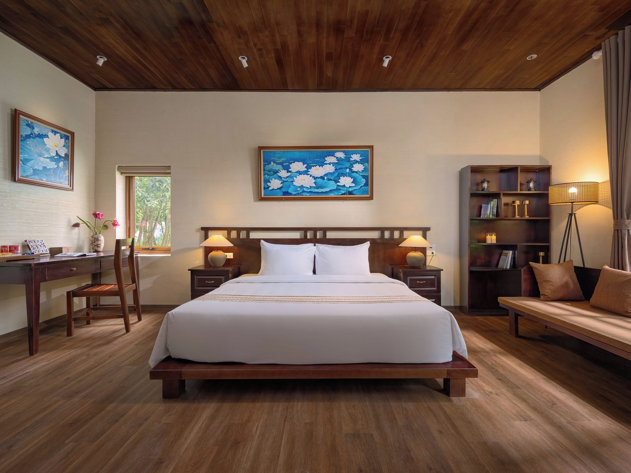 Habitación de hotel con una cama grande, cabecera de madera, dos mesitas de noche con lámparas, un escritorio con una silla, una estantería y una ventana con vistas a zonas verdes.