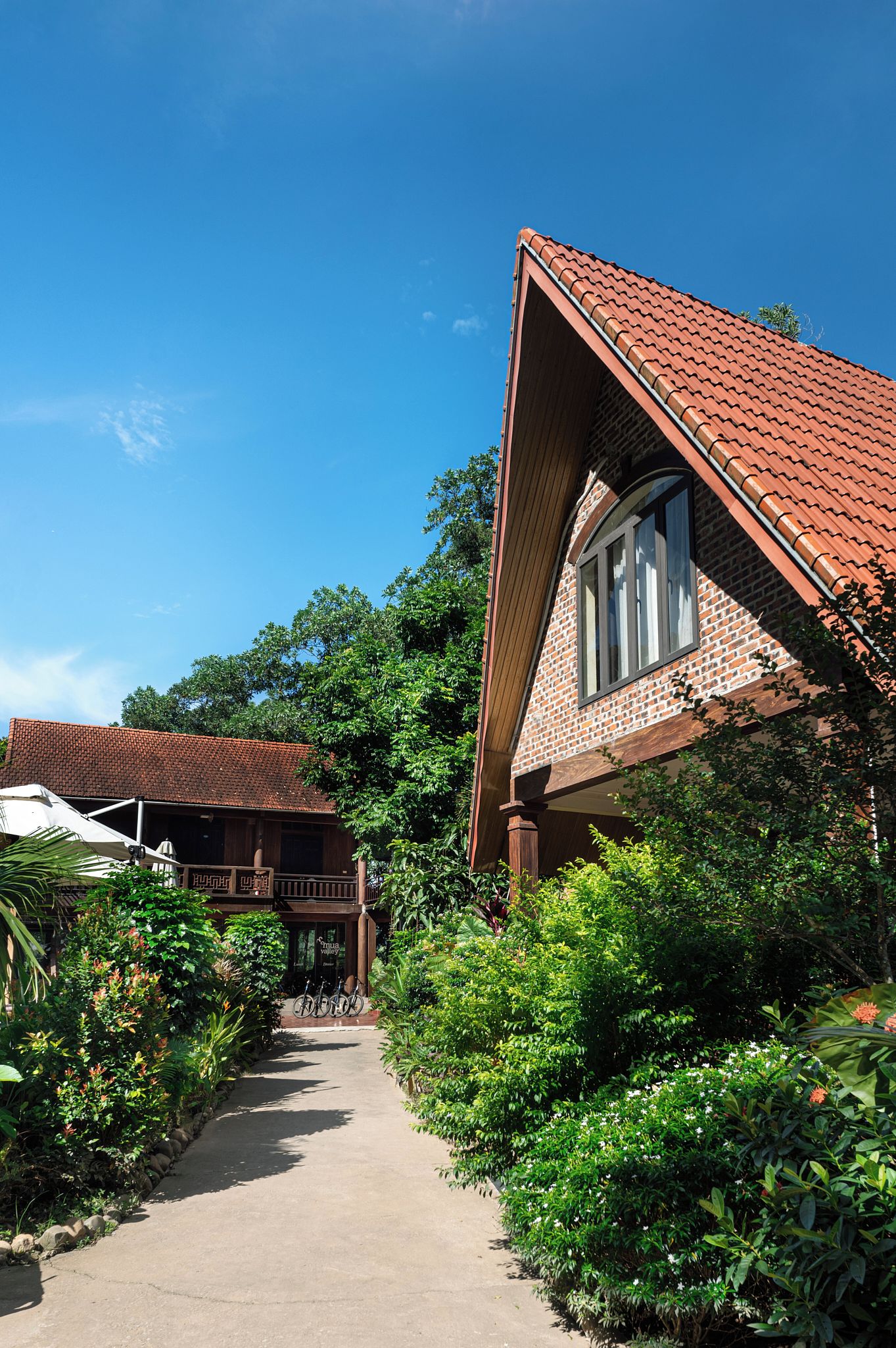 A pathway leading to a building with a red-tiled roof and brick walls, surrounded by lush greenery.