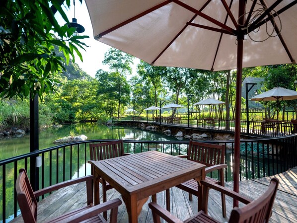 Une terrasse en bois, agrémentée d’une table et de chaises, entourée d’une rambarde en métal, et offrant une vue sur un étang paisible, avec des arbres et un pont en arrière-plan.