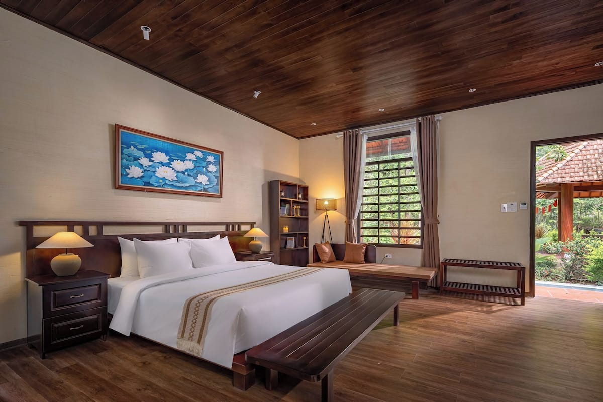 A spacious bedroom with a large bed, wooden headboard, bedside tables, a bench, and a view of a lush garden through a large window.