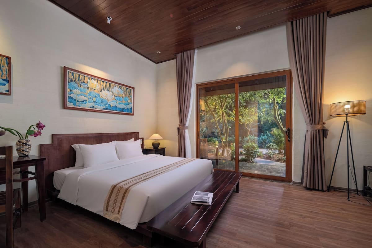 A hotel room with a wooden ceiling, a large bed with white bedding, a bench, a bedside table with a lamp, a painting on the wall, and a view of greenery through a sliding door.
