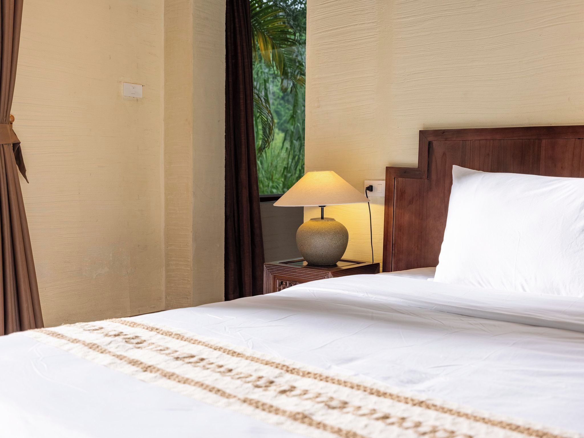 Una cama bien hecha con edredón blanco y manta color beige, una mesita de noche de madera con una lámpara, y una ventana con vistas a zonas verdes.