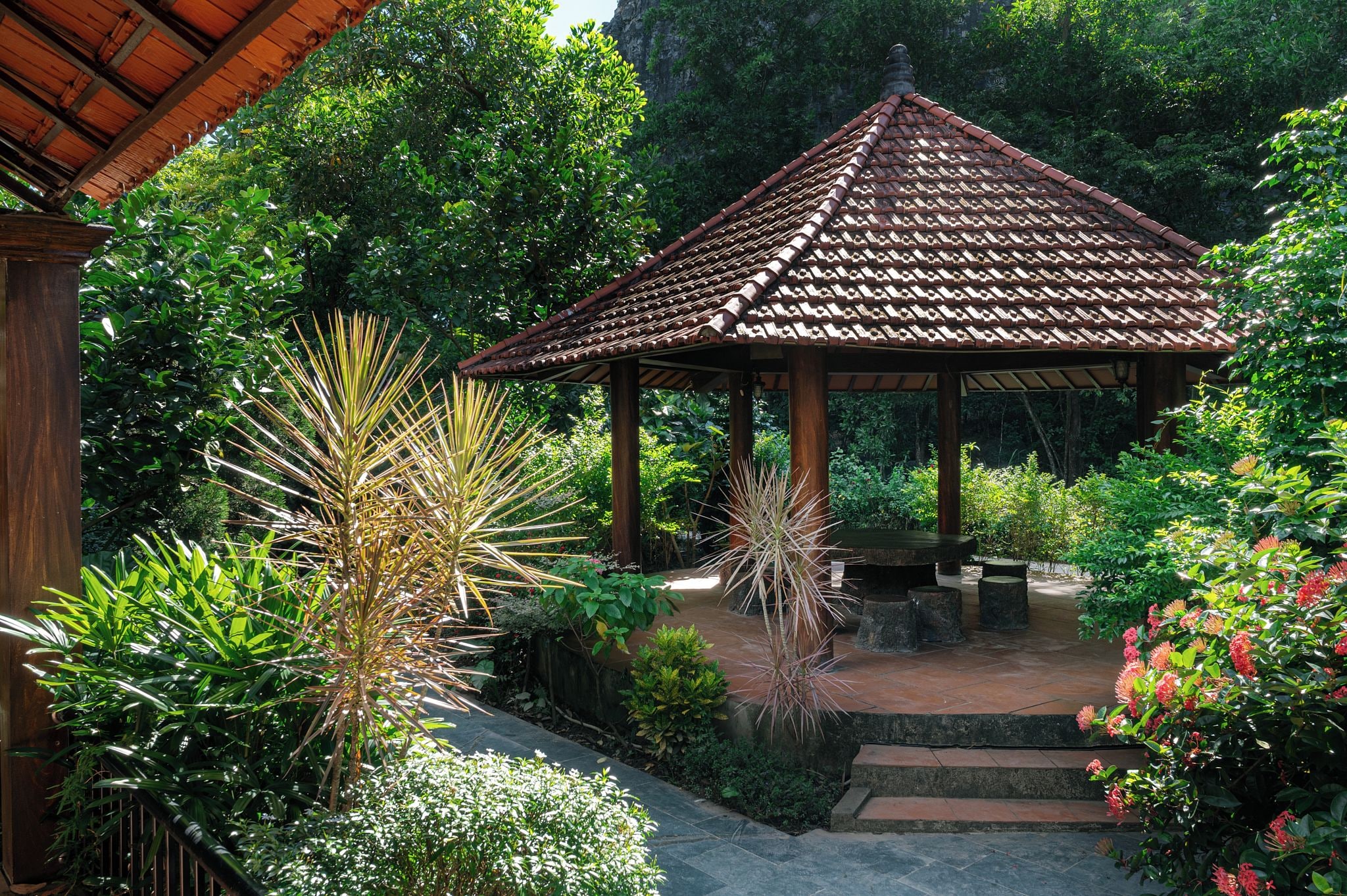 A gazebo with a tiled roof surrounded by lush greenery and a stone pathway.
