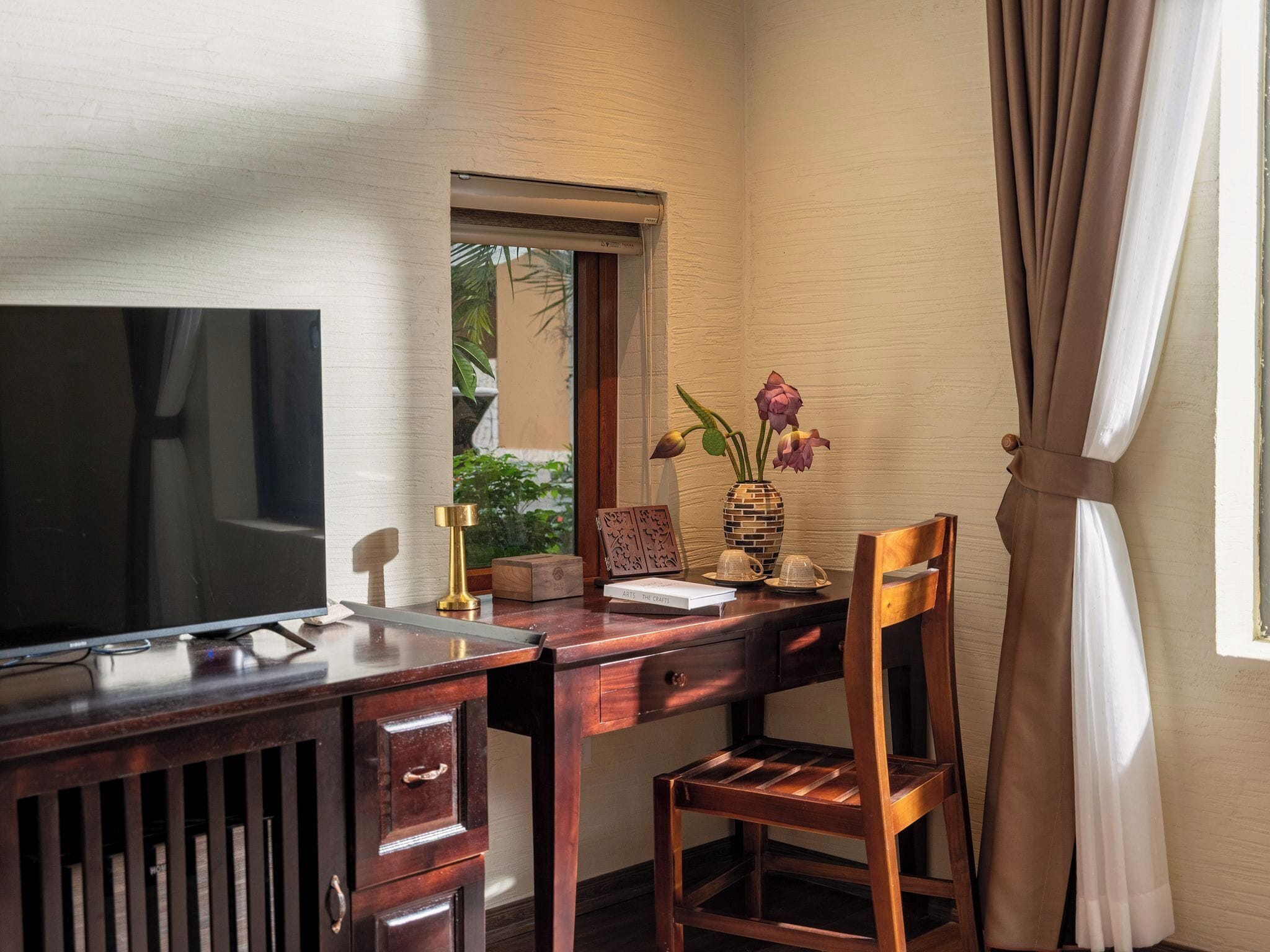 A wooden desk with a TV, a vase with flowers, and a window with curtains.