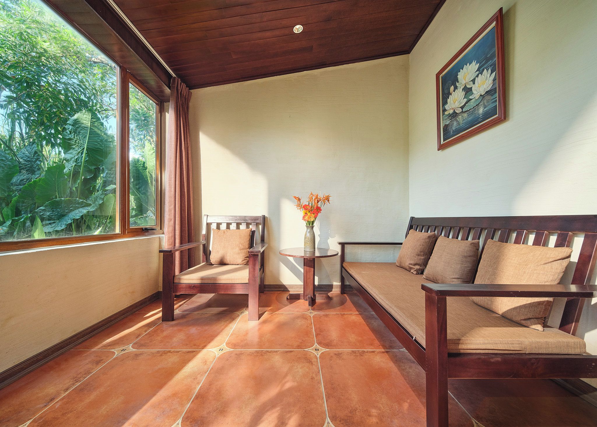 Una habitación con techo de madera, una ventana con cortinas, un sofá, una silla, una mesita con un jarrón y un cuadro en la pared.