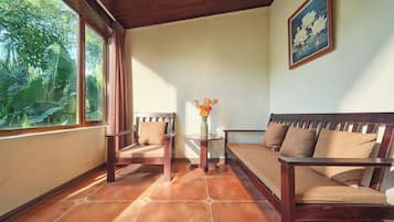 Una habitación con techo de madera, una ventana con cortinas, un sofá, una silla, una mesita con un jarrón y un cuadro en la pared.