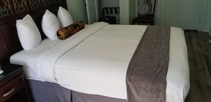 Un lit bien fait, avec du linge de lit blanc et un plaid marron, une table de chevet en bois avec un tiroir, et une chaise à l’arrière-plan.