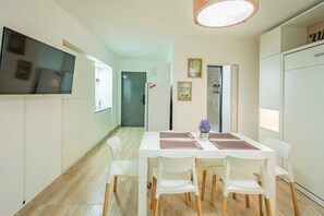 A modern kitchen with white cabinetry, a dining table with chairs, a flat-screen TV mounted on the wall, and a ceiling light fixture.