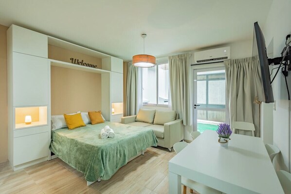 A hotel room with a bed, a sofa, a dining table, a TV, and a window with curtains.