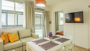 Ett modernt vardagsrum med en soffa, ett matbord med stolar, en platt-TV som är monterad på väggen och ett stort fönster med gardiner.