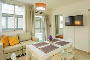A modern living room with a sofa, a dining table with chairs, a flat-screen TV mounted on the wall, and a large window with curtains.