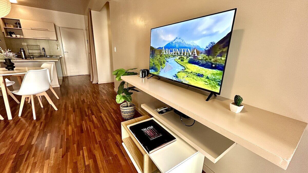 Una sala moderna con un televisor de pantalla plana mostrando una imagen panorámica de Argentina, un comedor con sillas blancas y una mesa de madera, y una cocina al fondo.