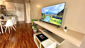 Een moderne woonkamer met een flatscreen televisie waarop een pittoresk beeld van Argentinië te zien is, een eethoek met witte stoelen en een houten tafel, en een keuken op de achtergrond.
