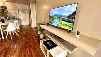 Una sala moderna con un televisor de pantalla plana mostrando una imagen panorámica de Argentina, un comedor con sillas blancas y una mesa de madera, y una cocina al fondo.