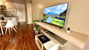A modern living room with a flat-screen TV displaying a scenic image of Argentina, a dining area with white chairs and a wooden table, and a kitchen in the background.