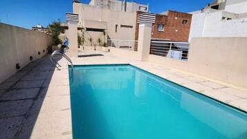 Una piscina en la azotea con un cielo azul despejado, rodeada de edificios.