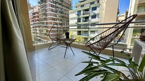 A balcony with two wicker chairs and a small table, overlooking a residential area with multi-story buildings.
