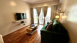A living room with a flat-screen TV, a green sofa, a wooden coffee table, and a balcony with curtains.