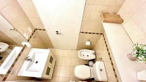Een badkamer met twee wastafels, een toilet en een bidet. Er is een handdoekhouder en een plant aan de muur.