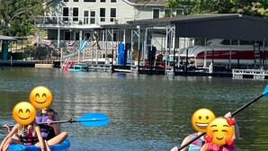 Deux personnes se trouvent dans un petit bateau gonflable bleu sur une étendue d’eau calme. On aperçoit un quai et une maison en arrière-plan.