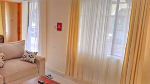 Ein Wohnzimmer mit einem beigefarbenen Sofa, gemusterten Kissen, einer Fernbedienung auf einem Holztisch und durchsichtigen Vorhängen an den Fenstern.