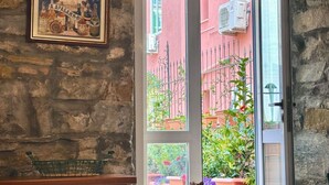 A room with a stone wall, a framed poster, a window with a view of a pink building, and a metal basket on the floor. - Casa Camelie, with a spacious terrace (Trieste)