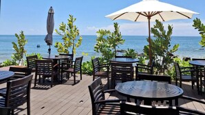 An outdoor patio with wooden tables and chairs, a white umbrella, and a view of the ocean. - Pandora Beach Mui Ne Retreat (Mui Ne)