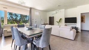 A modern living room with a dining area, white sofas, a flat-screen TV, and a large window offering a view of trees.