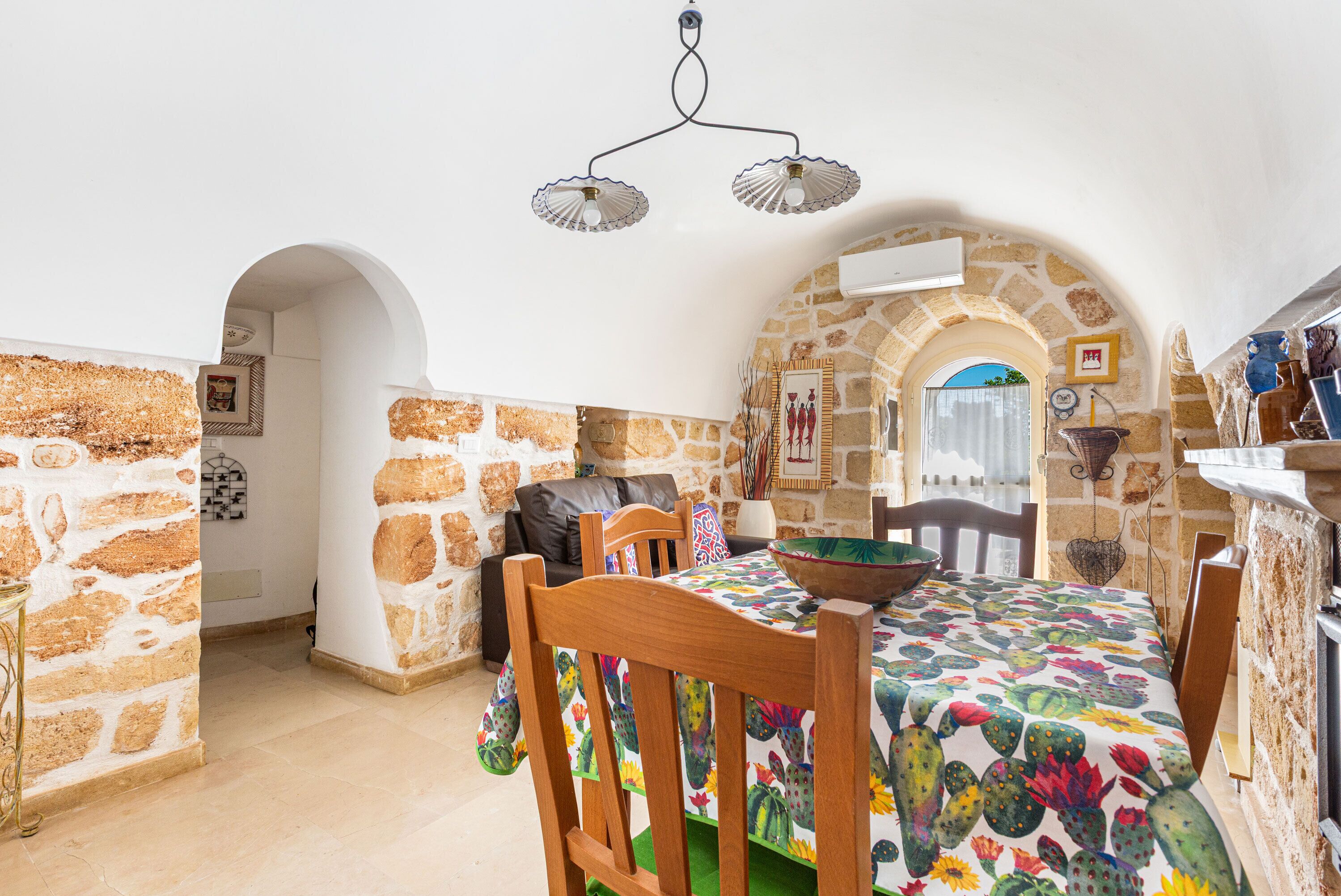 A dining area with a table set for two, a wooden chair, and a bowl on the table. The room features stone walls and a ceiling fan.