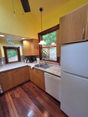 A kitchen with wooden cabinets, a stainless steel sink, a dishwasher, and a window with a view of a wooden deck and greenery. - The Cozy Camel (Marshall)