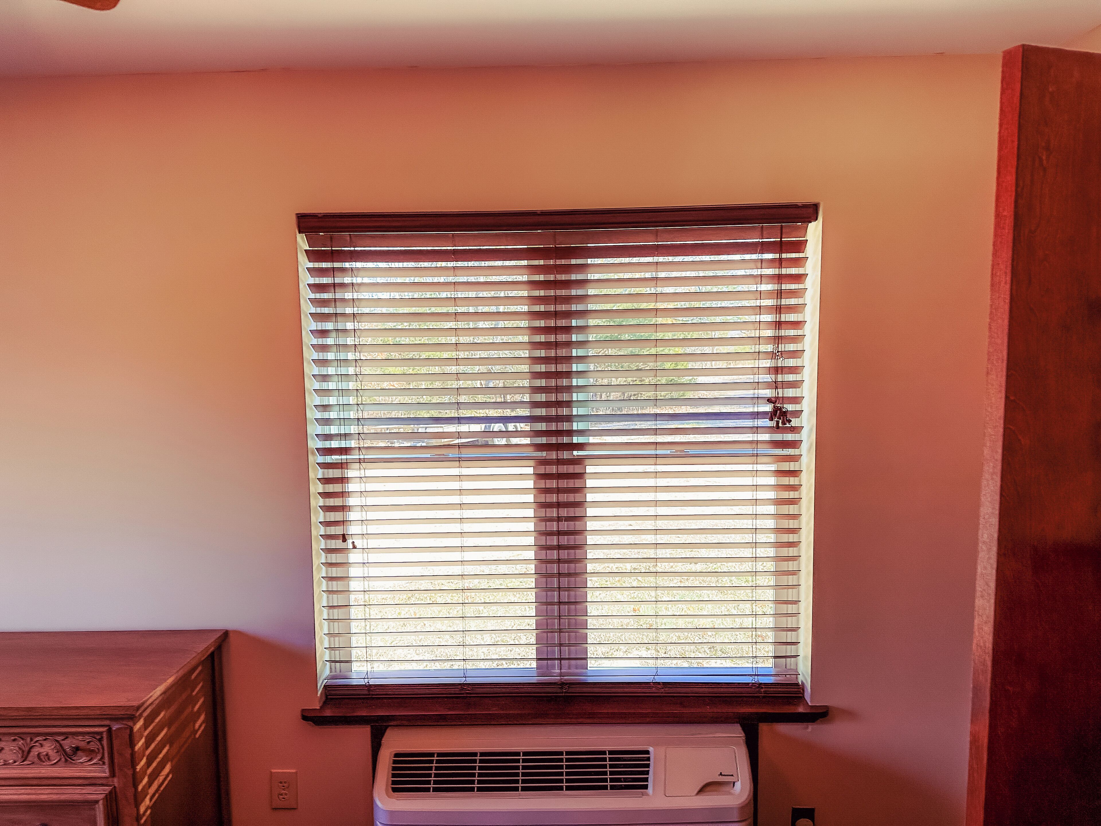 Une fenêtre avec des stores horizontaux, un climatiseur fixé au mur et un meuble en bois.