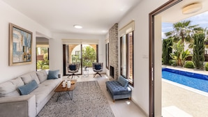 Una sala moderna con un sofá, sillones, una mesa de centro y una chimenea. Hay un ventanal con vista a una piscina y palmeras.