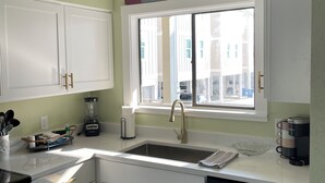 A modern kitchen with white cabinets, a built-in dishwasher, a window with a view of buildings, and a 'SEASIDE' sign above the window.