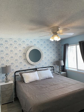 A bedroom with a bed, two bedside tables, a ceiling fan, a round mirror, and a window with curtains.