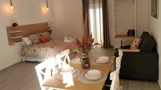 A cozy bedroom with a bed, bedside table, and a nightstand. There is a dining area with a table set for two, a chair, and a vase with flowers. - Antonio Parra II Tourist Apartments (Jerez de la Frontera)