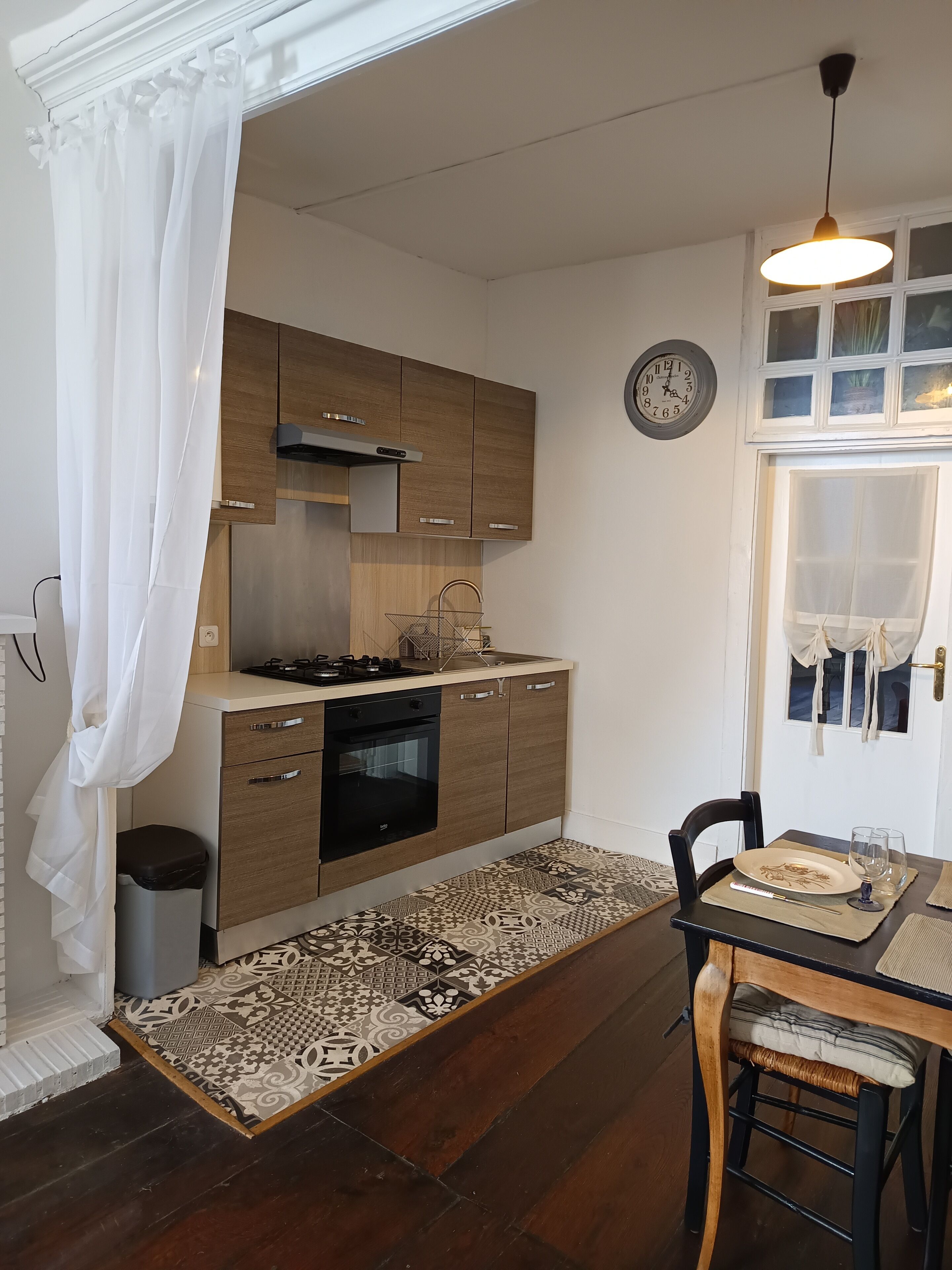 Une cuisine de taille modeste, avec des meubles en bois, une cuisinière à gaz et un évier. On y trouve une table à manger avec des chaises et un tapis à motifs.