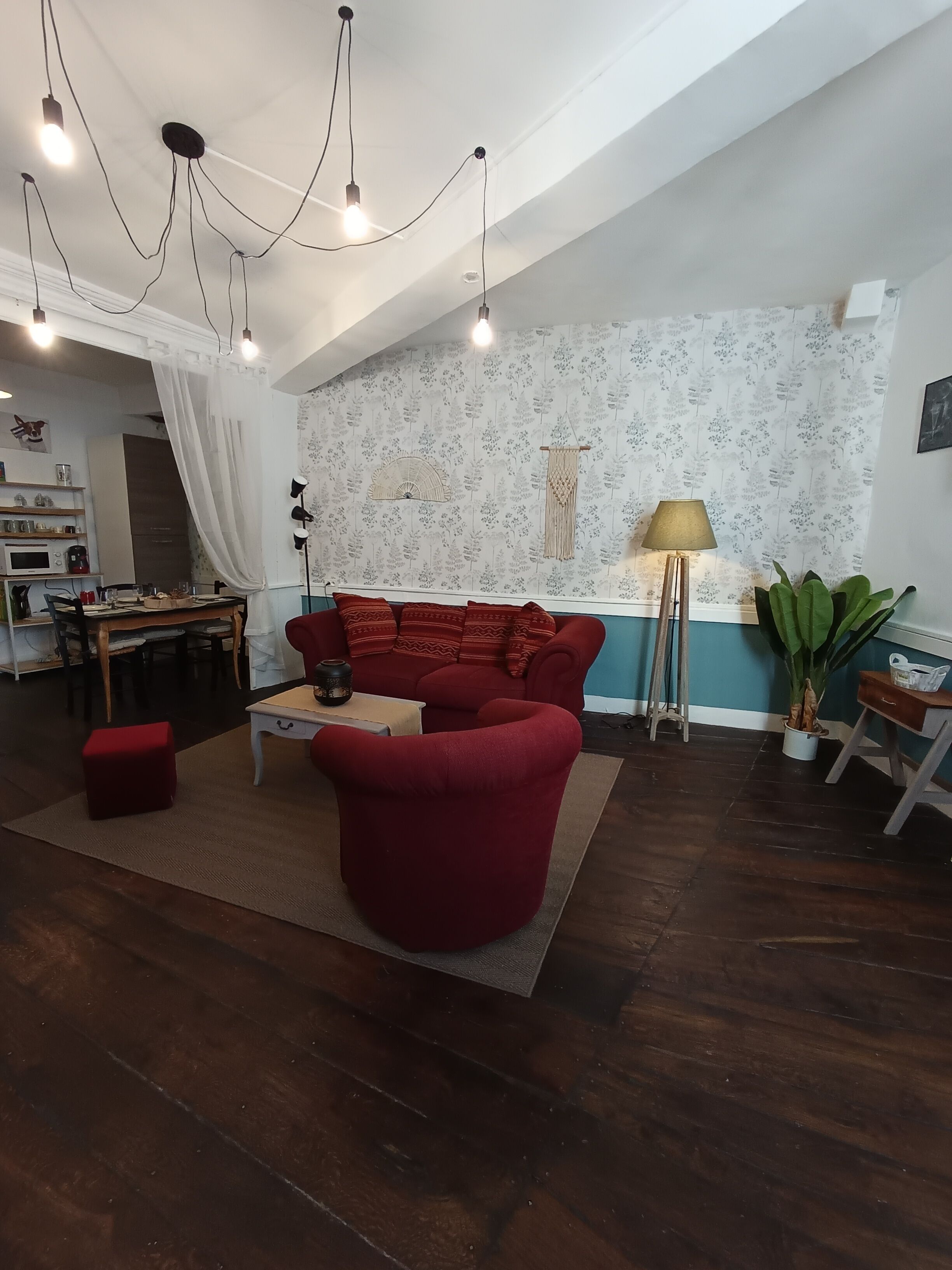 Un salon chaleureux avec un canapé rouge, une table basse et un coin repas comprenant une table et des chaises. On y trouve une lampe, une plante et un mur orné d’un motif floral.