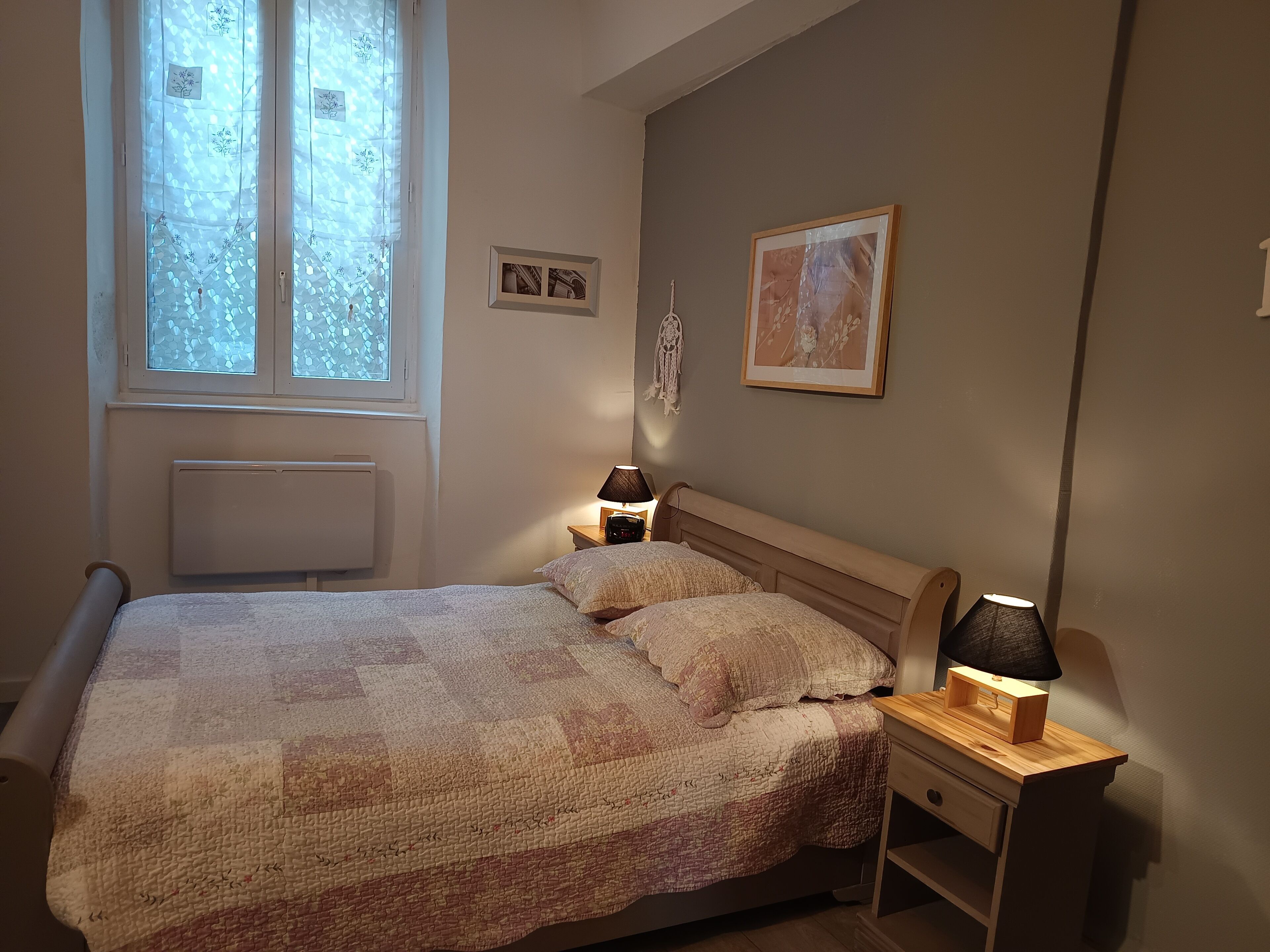 A bedroom with a bed, bedside lamps, a window with lace curtains, and wall decorations.