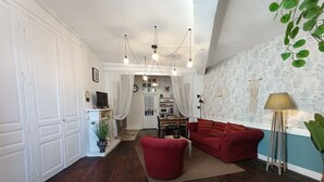 Un salón con un sofá rojo, una mesa de centro y un televisor sobre un mueble. Hay una chimenea con una planta a su lado. El piso es de madera y el papel tapiz tiene un diseño.