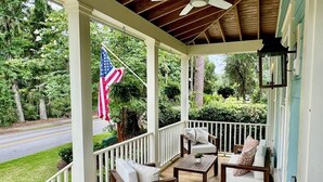 A covered porch with wooden flooring, white railings, and a ceiling fan. There are two white sofas with cushions and a small table. An American flag is visible outside the porch. - Wharf Street Coastal Escape - 3BR Old Town Luxury (Bluffton)