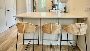 A modern kitchen with a central island, three wicker bar stools, pendant lighting, and white cabinetry. - Wharf Street Coastal Escape - 3BR Old Town Luxury (Bluffton)